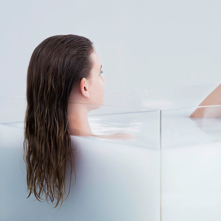 Kobieta z długimi włosami leży w szklanej wannie. Kampania marki Reverie, kosmetyki dostępne w butiku Beauty Rebel w Polsce.